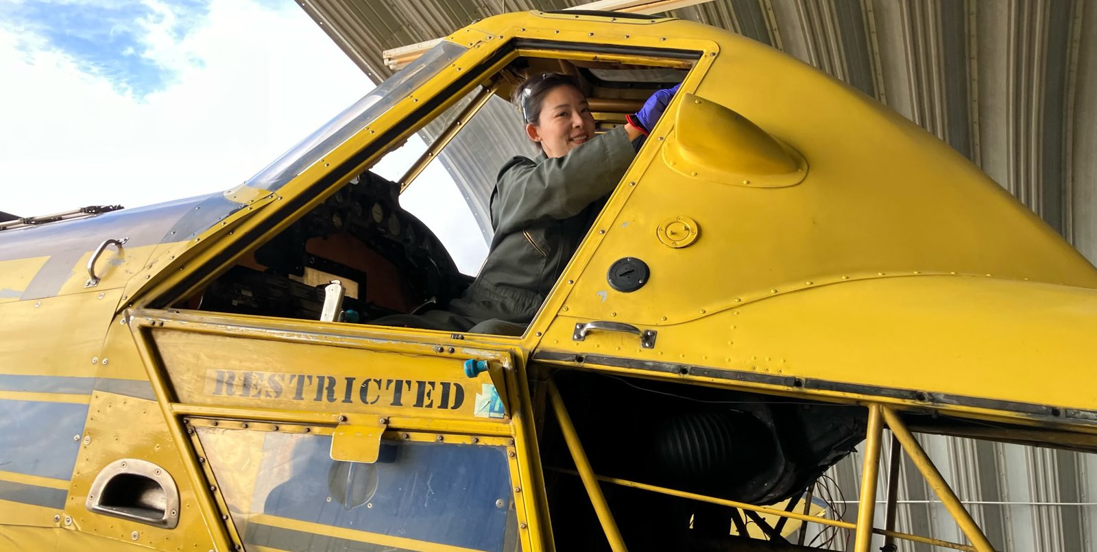 Person sitting in the cockpit of a yellow aircraft with "RESTRICTED" written on the side, under a metal roof.