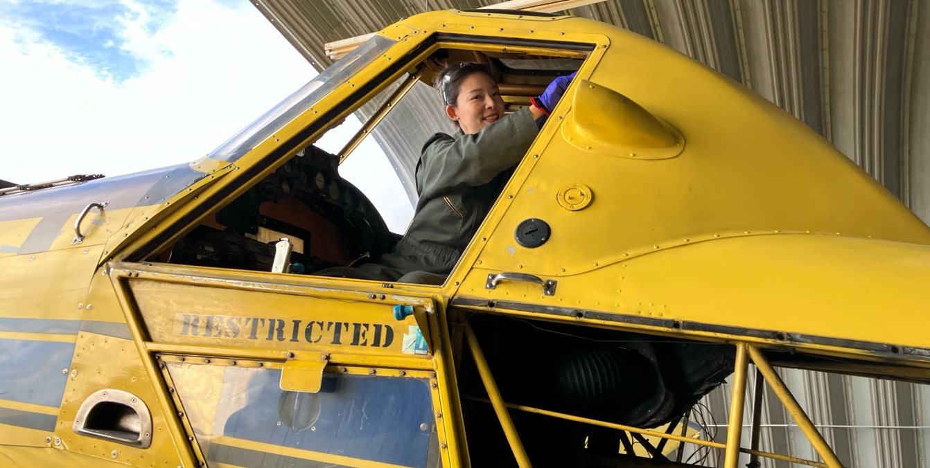 Person sitting in the cockpit of a yellow aircraft with "RESTRICTED" written on the side, under a metal roof.