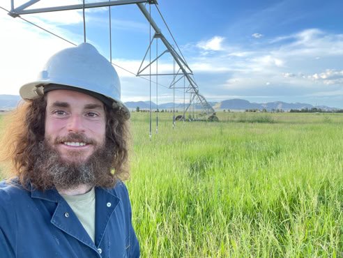 A person wearing a hard hat and blue jacket stands in a grassy field with an irrigation system overhead and a blue sky with clouds in the background.